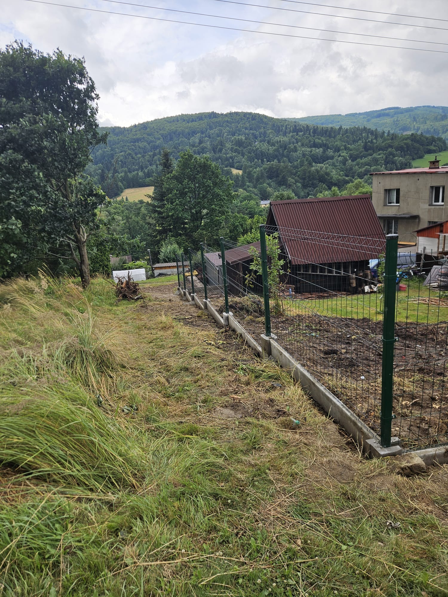 Montaż ogrodzenia panelowego na betonowej podmurówce, widok z góry na posesję z zaroślami i budynkiem w tle, teren pagórkowaty.