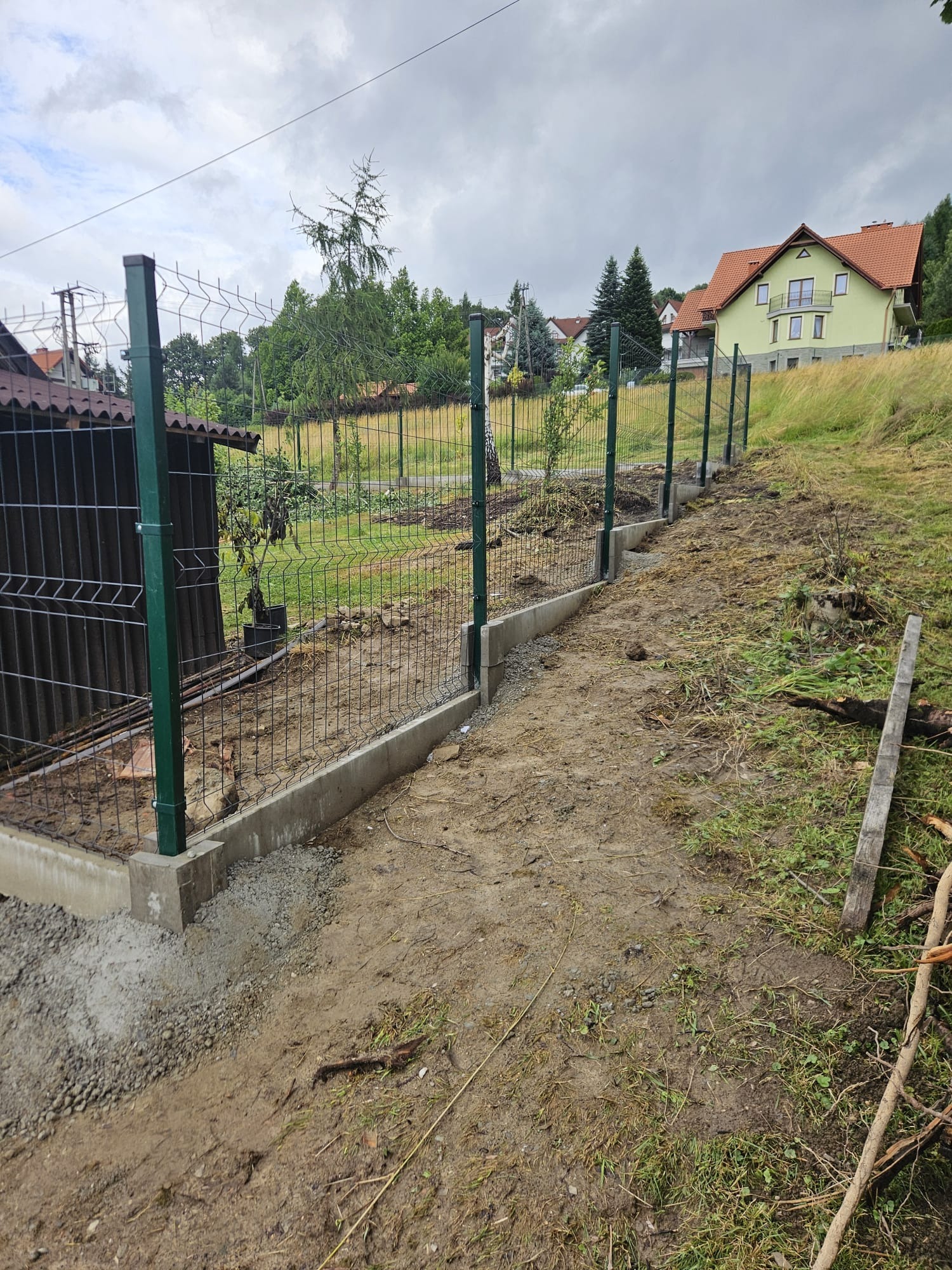 Montaż ogrodzenia panelowego z podmurówką betonową na nierównym terenie. Widoczne słupki w kolorze ciemnej zieleni i siatka w kolorze grafitowym, dom w tle.