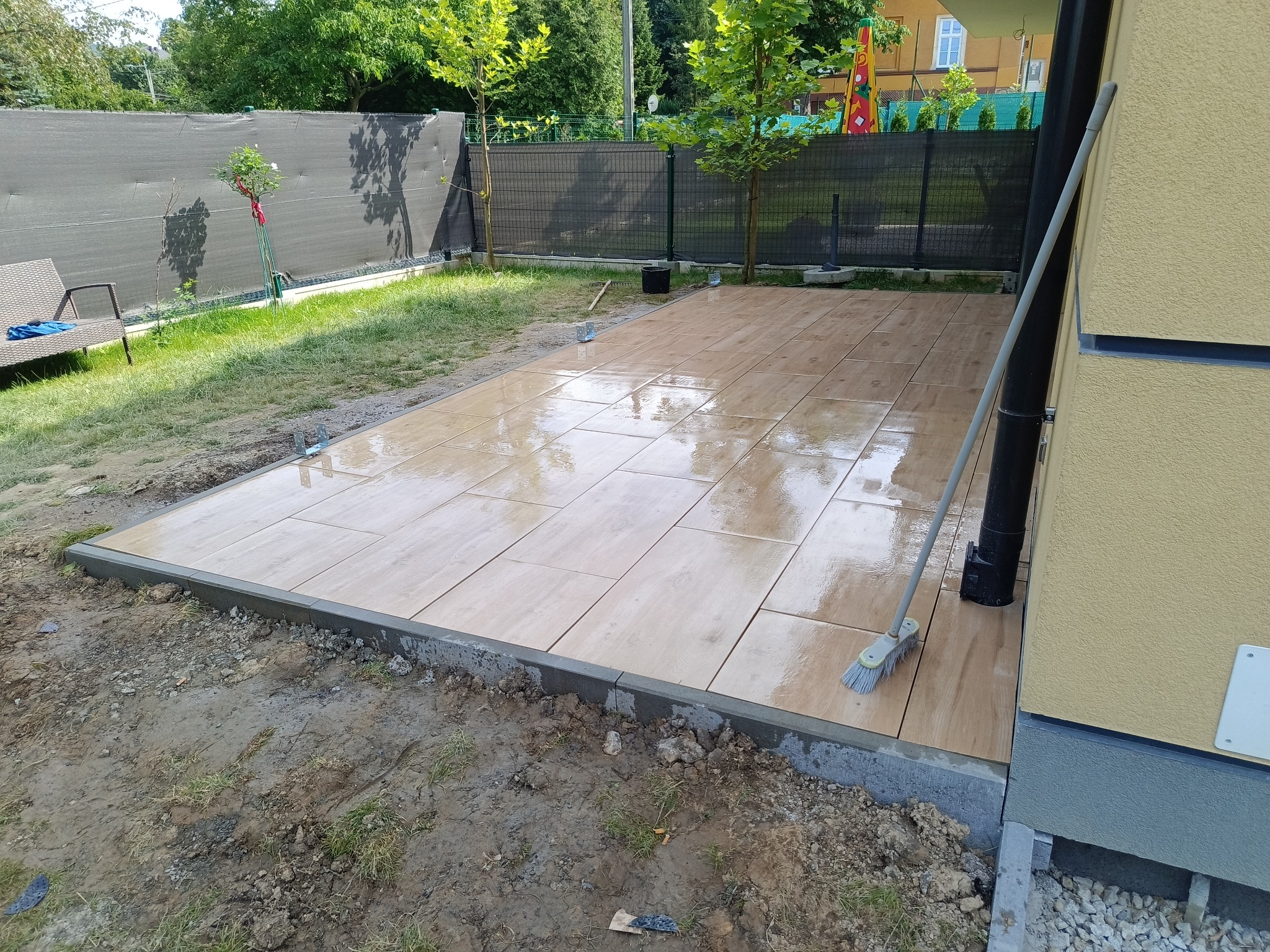 Ułożony taras z płytek ceramicznych imitujących drewno, obrzeża z szarej kostki betonowej, widoczny fragment domu i ogrodzenie panelowe.