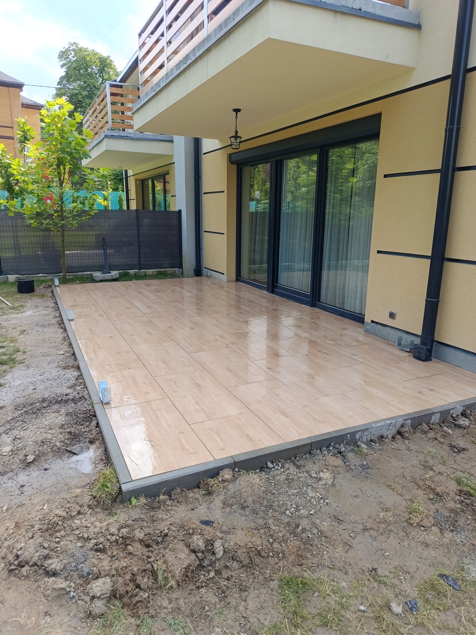 Nowoczesny taras z płytek imitujących drewno, wykończony szarą obwódką, przylegający do budynku z dużymi przesuwnymi drzwiami i balkonem na piętrze.