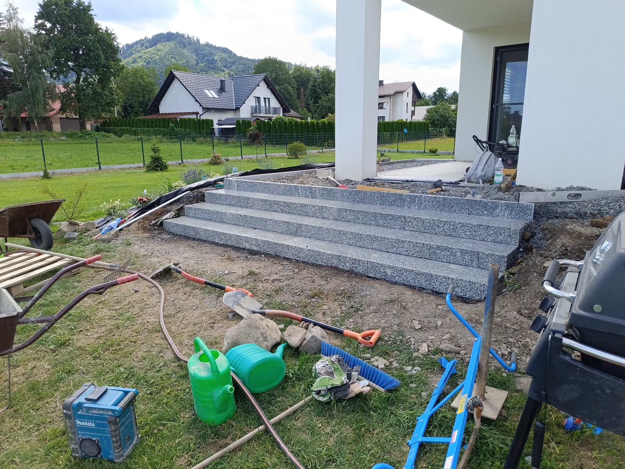 Granitowe schody wejściowe do nowoczesnego domu w trakcie montażu. W tle widoczne domy jednorodzinne i zieleń. Narzędzia i materiały porozrzucane wokół.