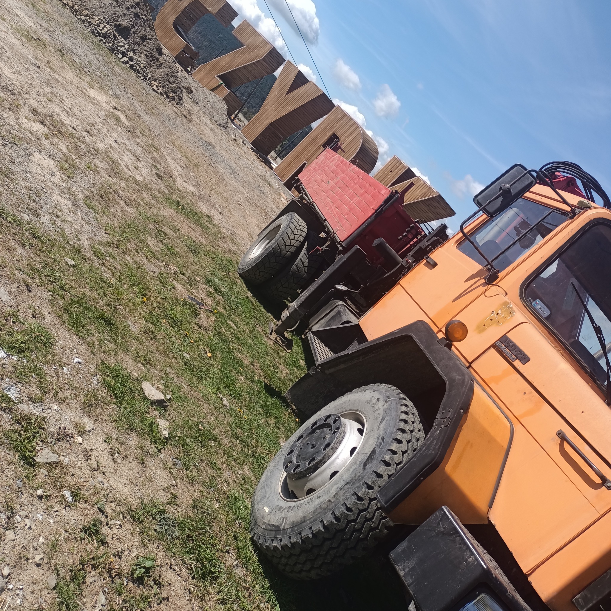 Pomarańczowa ciężarówka z czerwoną skrzynią ładunkową na ziemi, z drewnianą konstrukcją w tle, sfotografowana pod kątem, w słoneczny dzień.