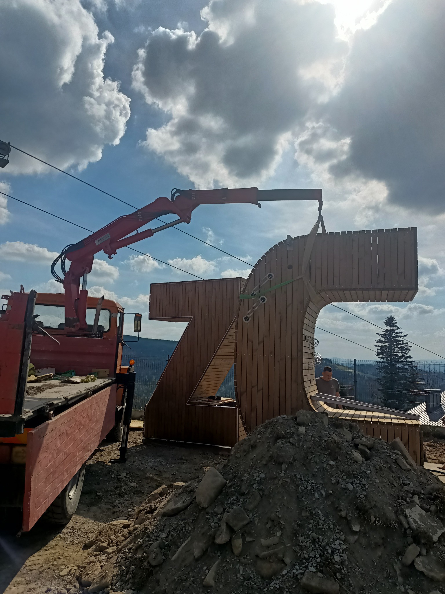 Montaż drewnianej konstrukcji w kształcie cyfry '70' za pomocą dźwigu samochodowego. W tle widoczne góry i błękitne niebo z chmurami.