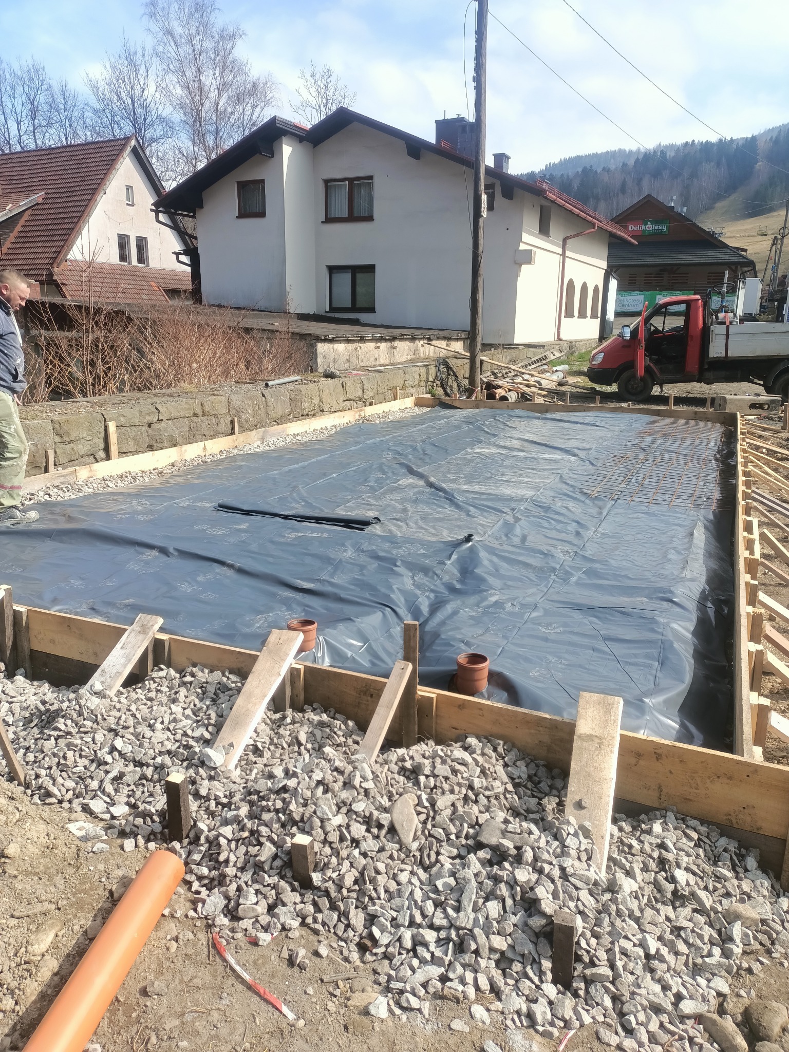 Przygotowanie podłoża: szalunek z desek, folia, zbrojenie i kamień. Widoczne budynki w tle i pracownik. Etap budowy z użyciem drewnianych elementów.