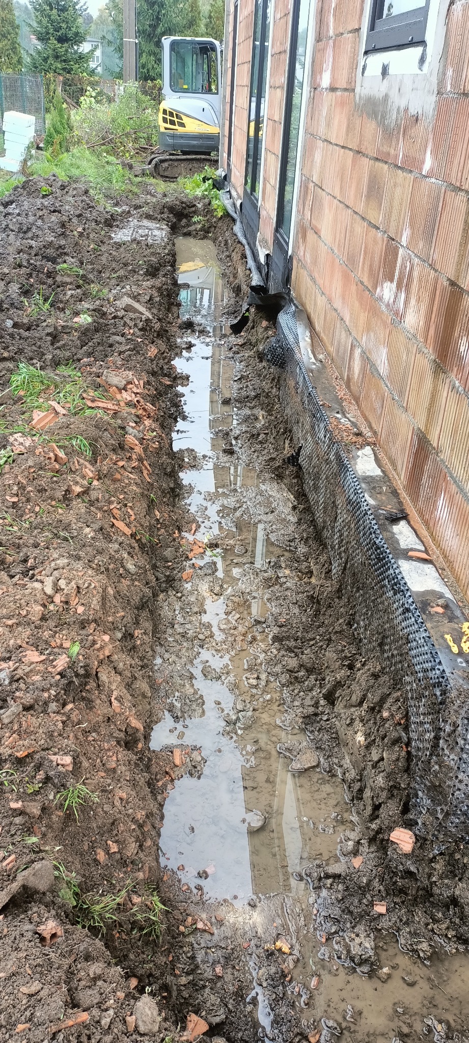 Wykop wzdłuż fundamentu budynku z widoczną folią kubełkową i stojącą wodą. W tle minikoparka. Prace ziemne przy nowym budynku w trakcie realizacji.