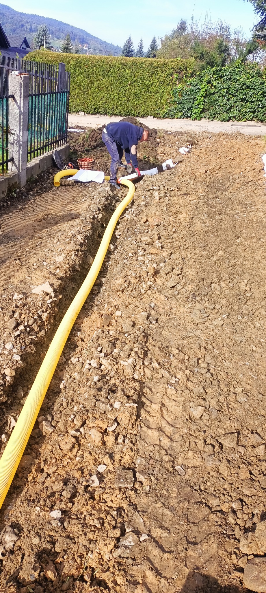 Żółta rura w wykopie, instalacja w ziemi. Prace ziemne przy ogrodzeniu, pracownik w odzieży roboczej. Widoczne narzędzia i materiały budowlane.