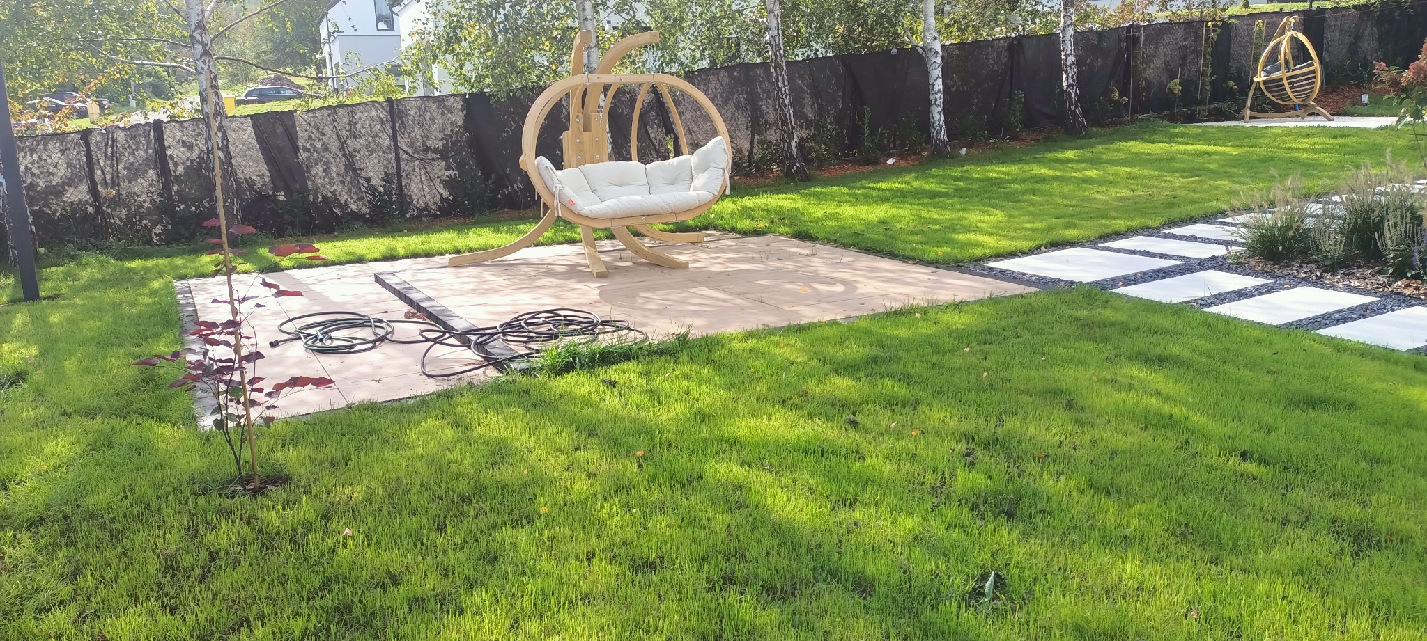 Ukończony taras z drewnianym fotelem wiszącym w kształcie kokonu, na tle zielonego trawnika i ogrodzenia z siatki maskującej. Widoczna ścieżka z płyt.