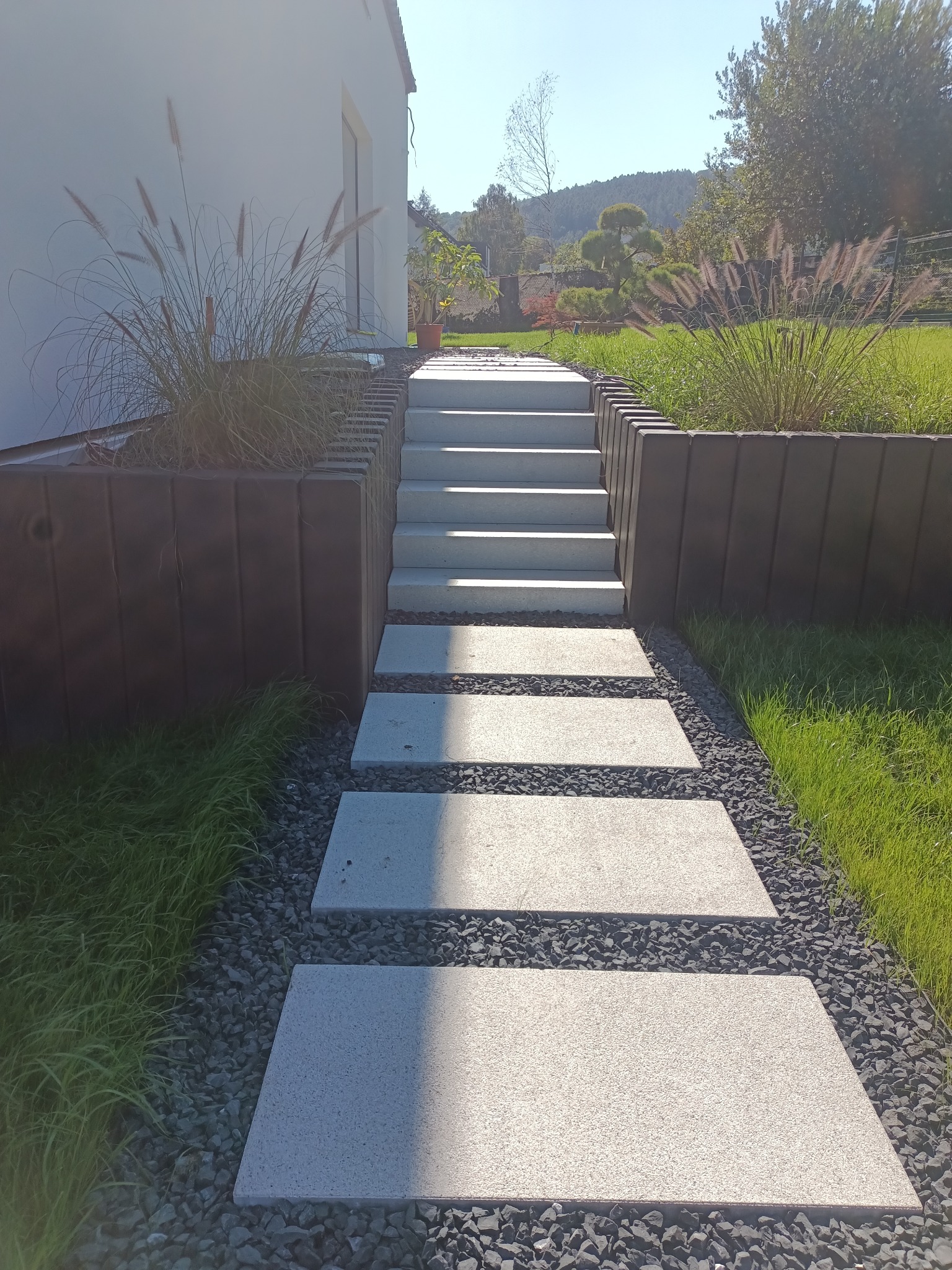 Nowoczesna ścieżka z płyt betonowych prowadząca do schodów, otoczona ozdobnym żwirem i zielenią. Widok na zadbany ogród z elementami małej architektury.