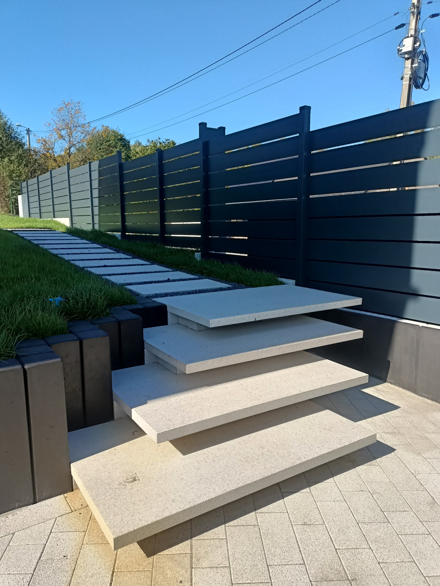 Nowoczesne ogrodzenie panelowe w kolorze antracytowym, schody z jasnego kamienia prowadzące w górę, trawnik z kamienną ścieżką. Słoneczny dzień.