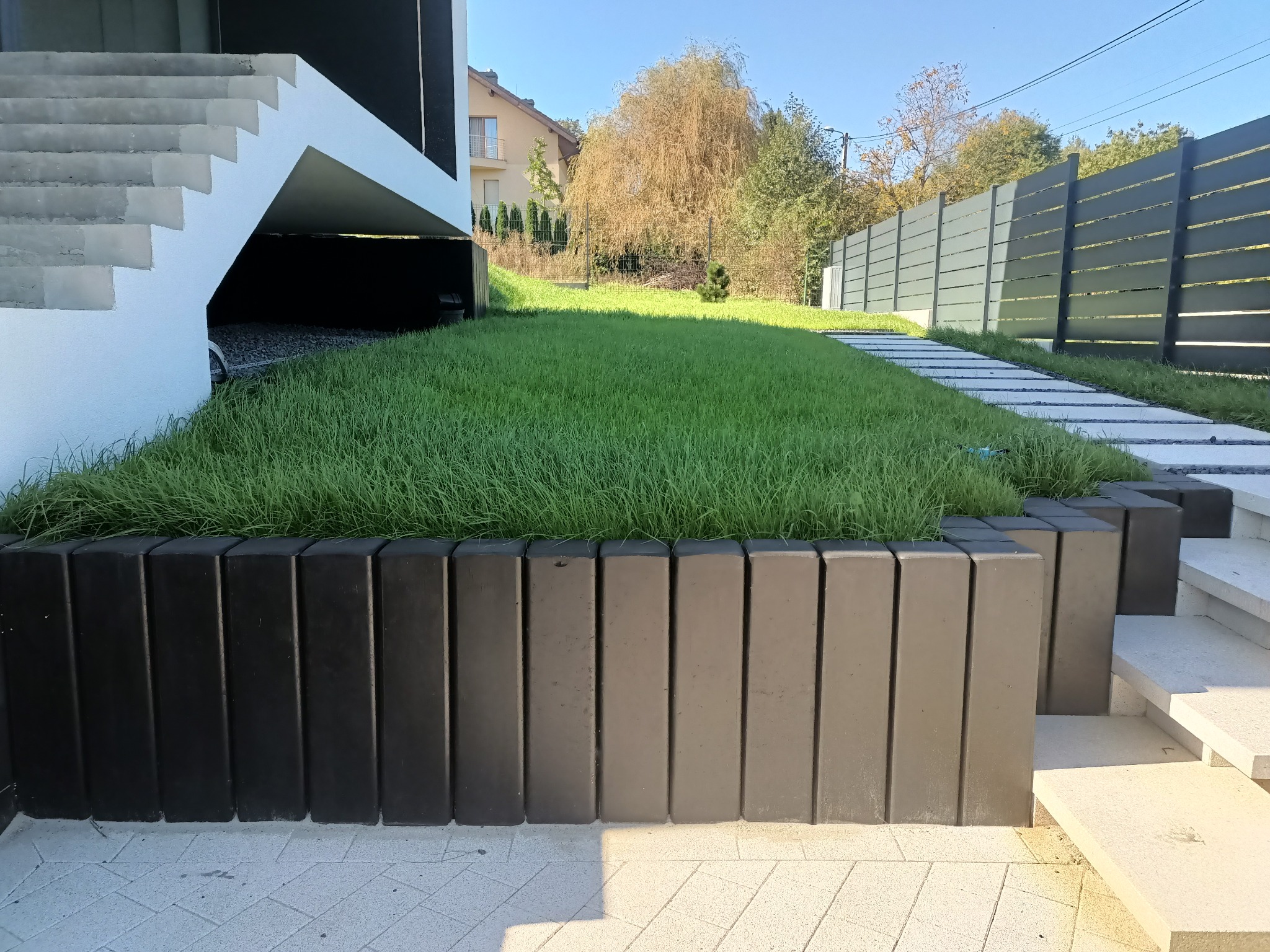 Nowoczesny ogród z geometryczną rabatą, czarnym obrzeżem i ścieżką z płyt. W tle widoczny dom i ogrodzenie panelowe. Całość skąpana w popołudniowym słońcu.