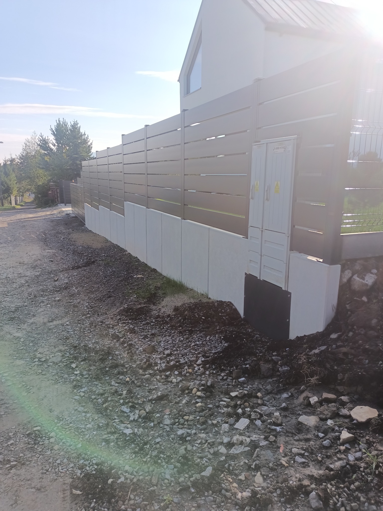 Nowoczesne ogrodzenie panelowe z poziomych listew, osadzone na betonowej podmurówce. Widoczne skrzynki elektryczne na ogrodzeniu i teren po budowie.