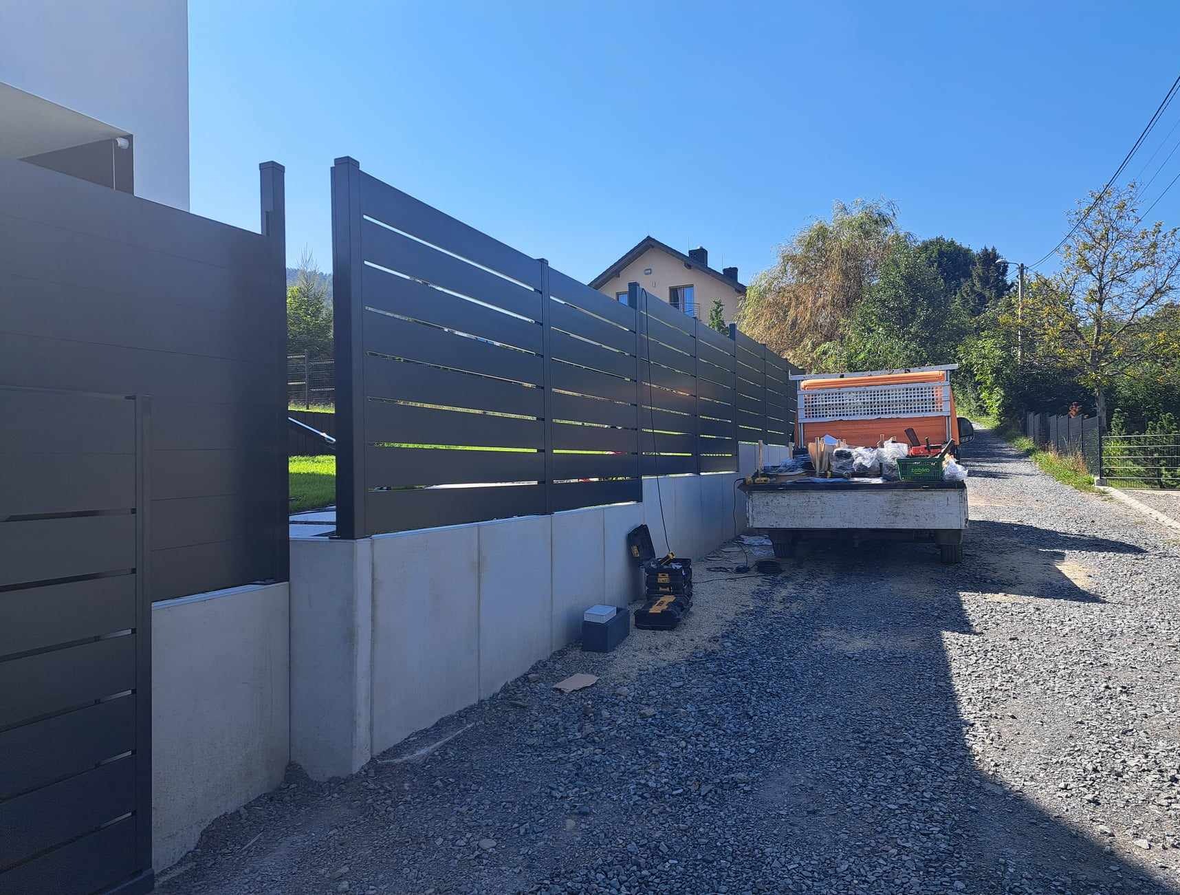 Nowoczesne ogrodzenie panelowe z poziomych profili, zamontowane na betonowym murku oporowym. W tle zaparkowany samochód dostawczy i dom jednorodzinny.