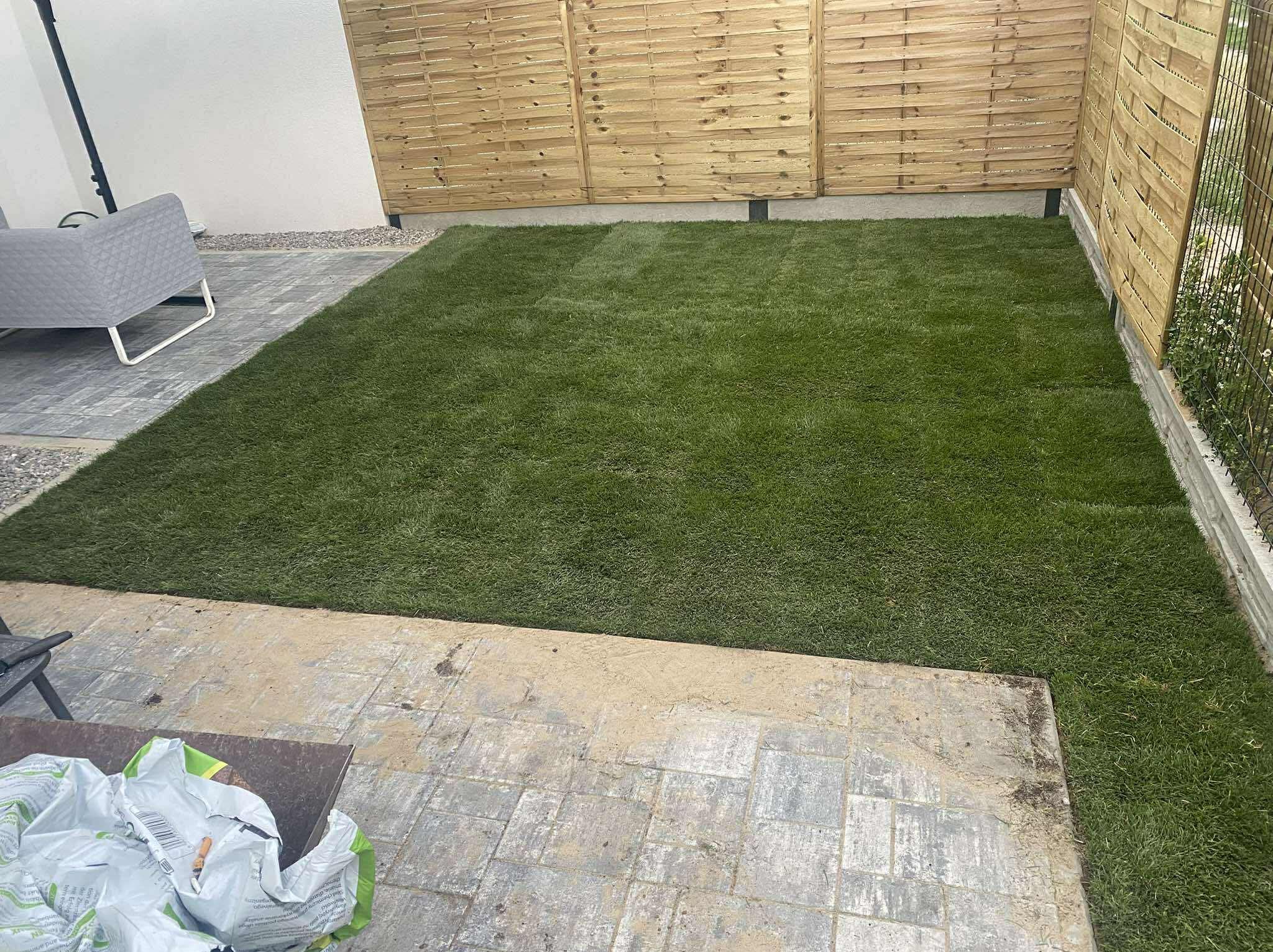 Świeżo ułożona, soczysta trawa z rolki w ogrodzie, widoczna z góry. Drewniane panele ogrodzeniowe i szara kostka brukowa tworzą schludne obramowanie.