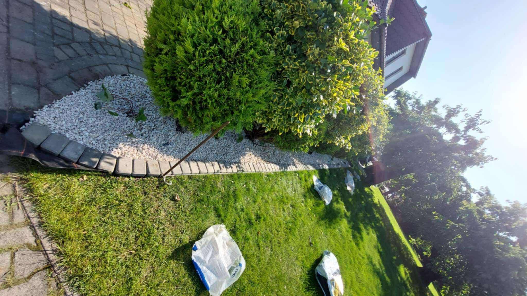 Aranżacja ogrodu z kamieniami i obrzeżami z kostki, widoczne worki z nawozem na trawniku. Ujęcie z boku, w tle zieleń i fragment budynku.