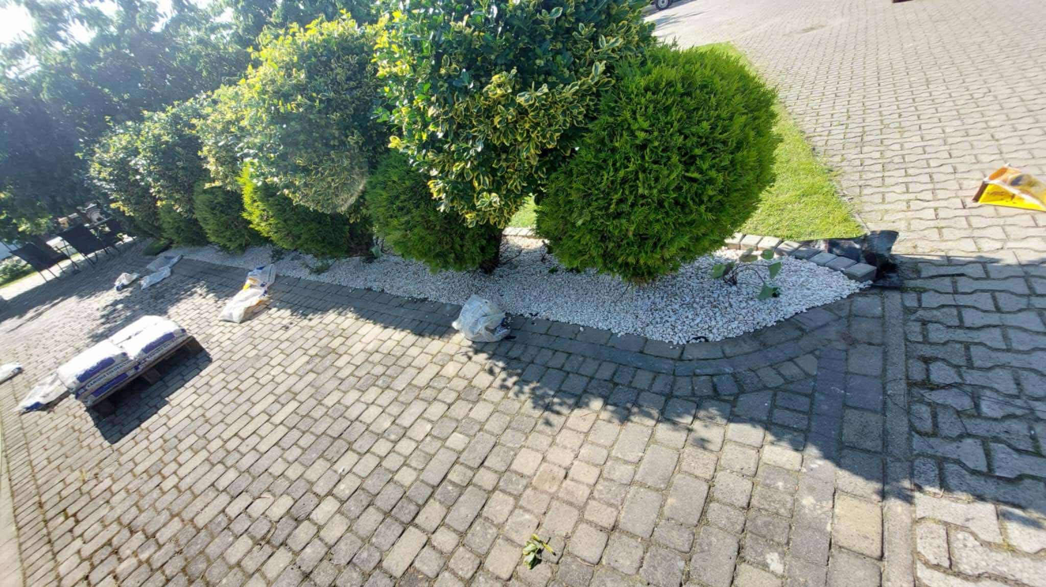 Formowane krzewy wzdłuż ścieżki z kostki brukowej, podsypane białym kamieniem. Worki z materiałem budowlanym obok. Widok z góry, oświetlenie słoneczne.