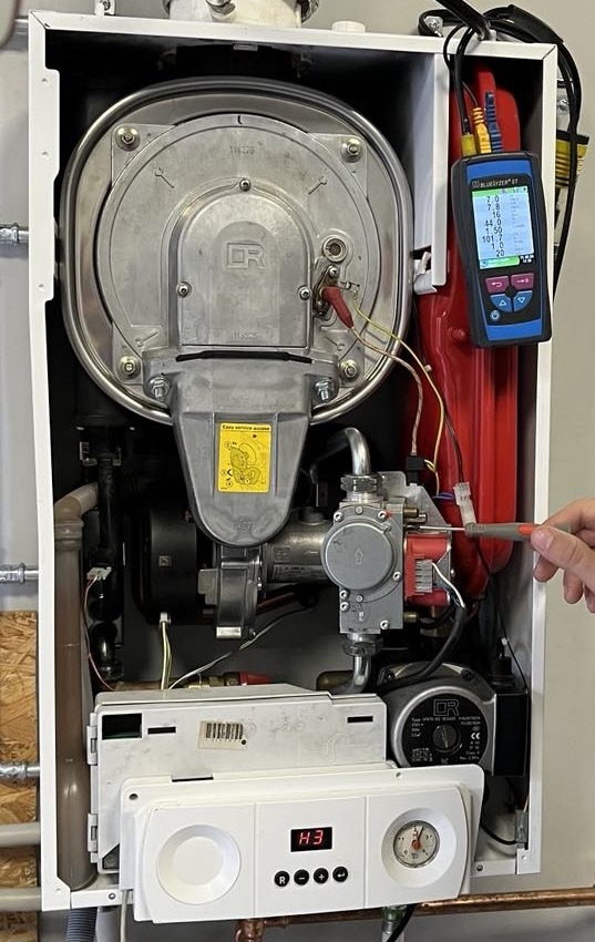 Serwis pieca gazowego: technik reguluje ustawienia, używając śrubokręta i analizatora spalin BlueLyzer STx. Widoczne elementy pieca i panel sterowania.