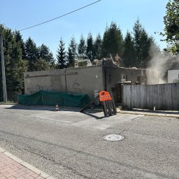 Pan Demolka - Rozbiórka niskiego budynku z graffiti na ścianie. Dwóch robotników na dachu w trakcie pracy, unoszący się pył. Na pierwszym planie ulica i elementy zabezpieczeń.