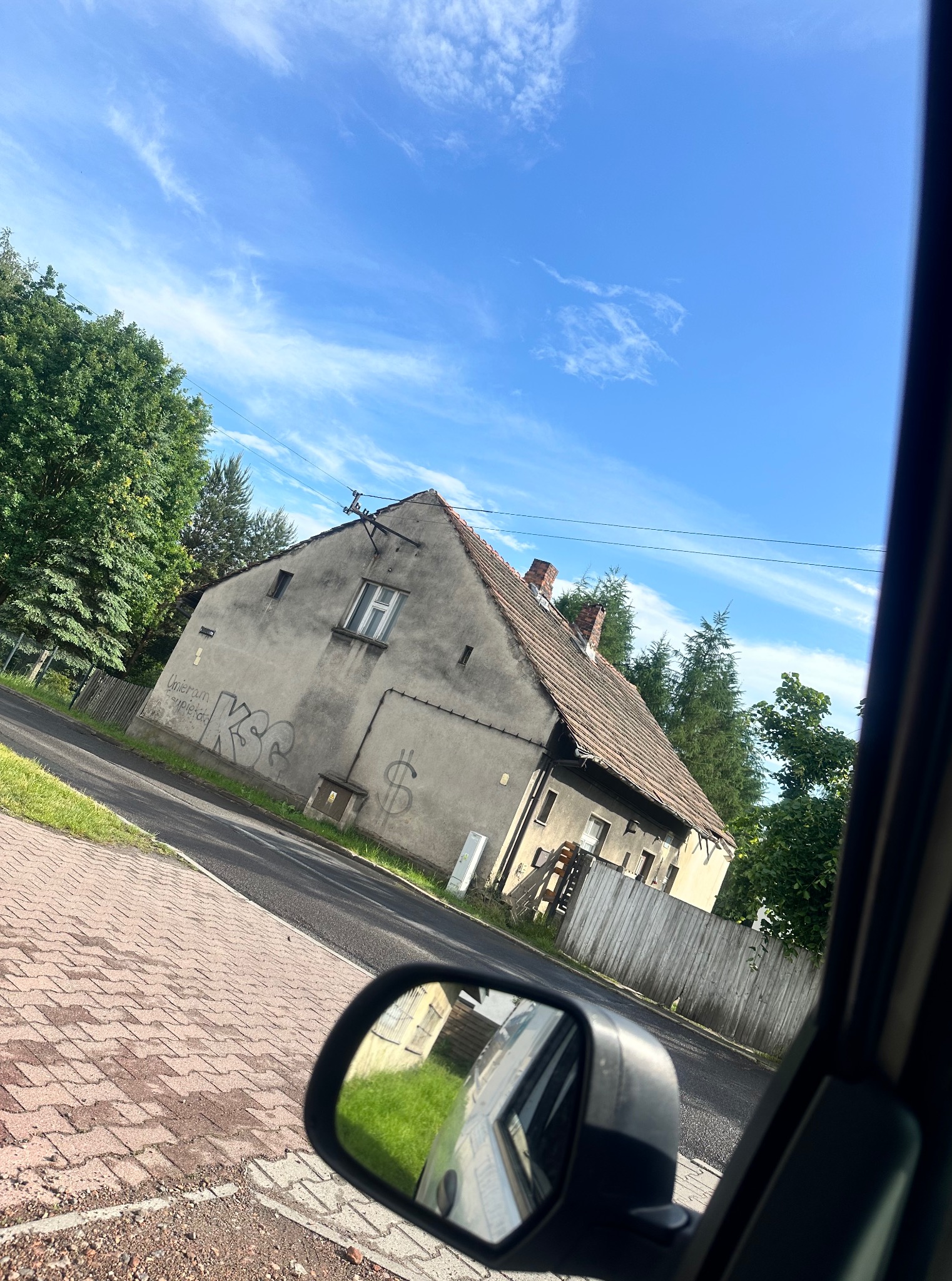 Szary, zaniedbany dom z graffiti i znakiem dolara na ścianie, widziany z wnętrza samochodu, z fragmentem lusterka bocznego i zielenią w tle.