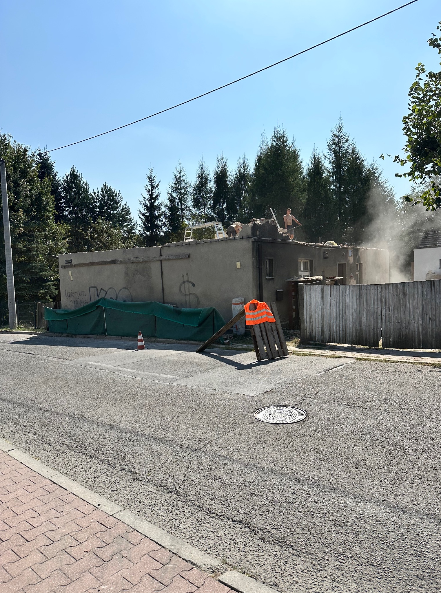 Rozbiórka niskiego budynku z graffiti na ścianie. Dwóch robotników na dachu w trakcie pracy, unoszący się pył. Na pierwszym planie ulica i elementy zabezpieczeń.