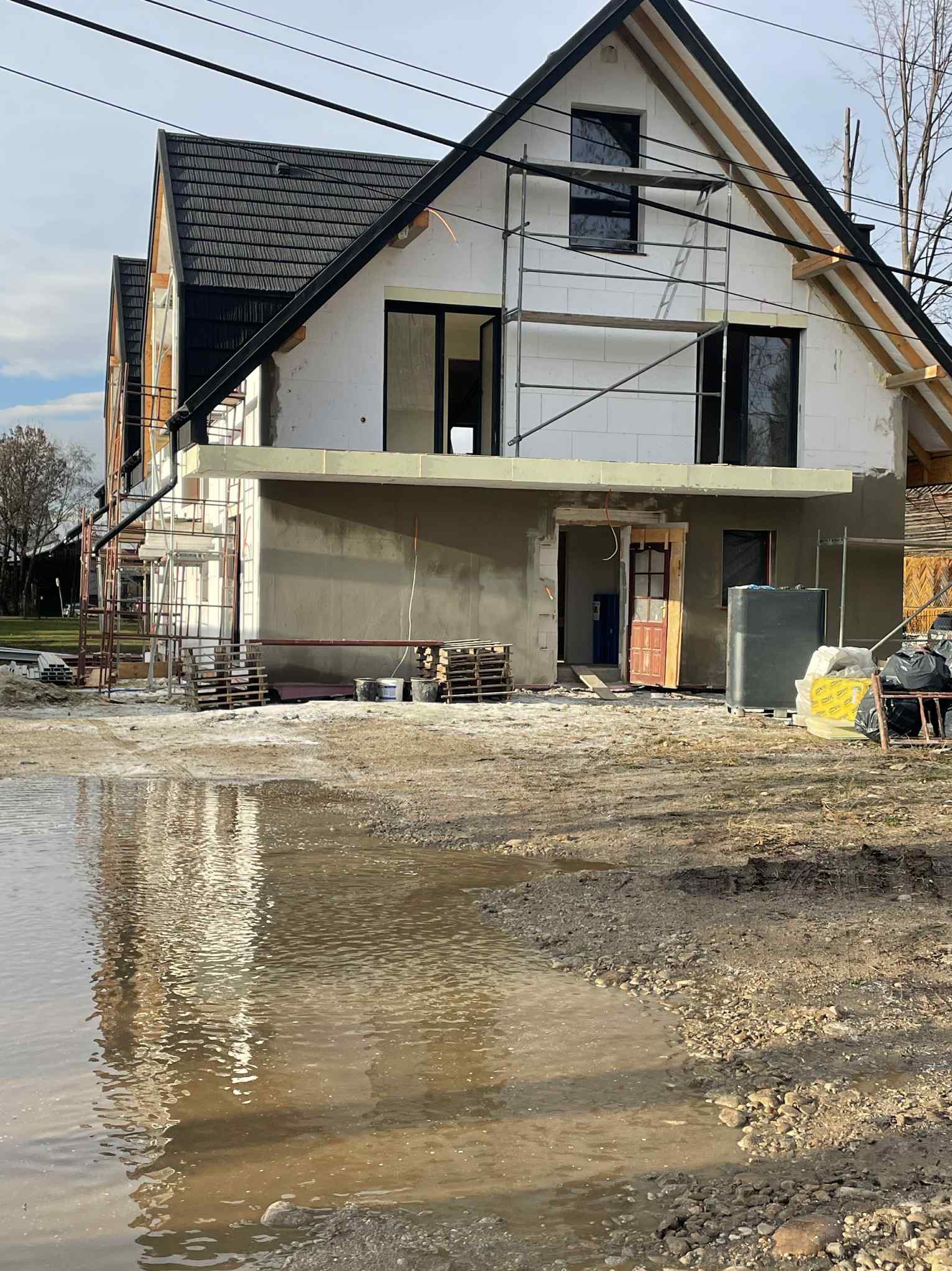 Dom w trakcie ocieplania elewacji, widoczne rusztowania, styropian i częściowo otynkowana ściana. Na pierwszym planie kałuża.