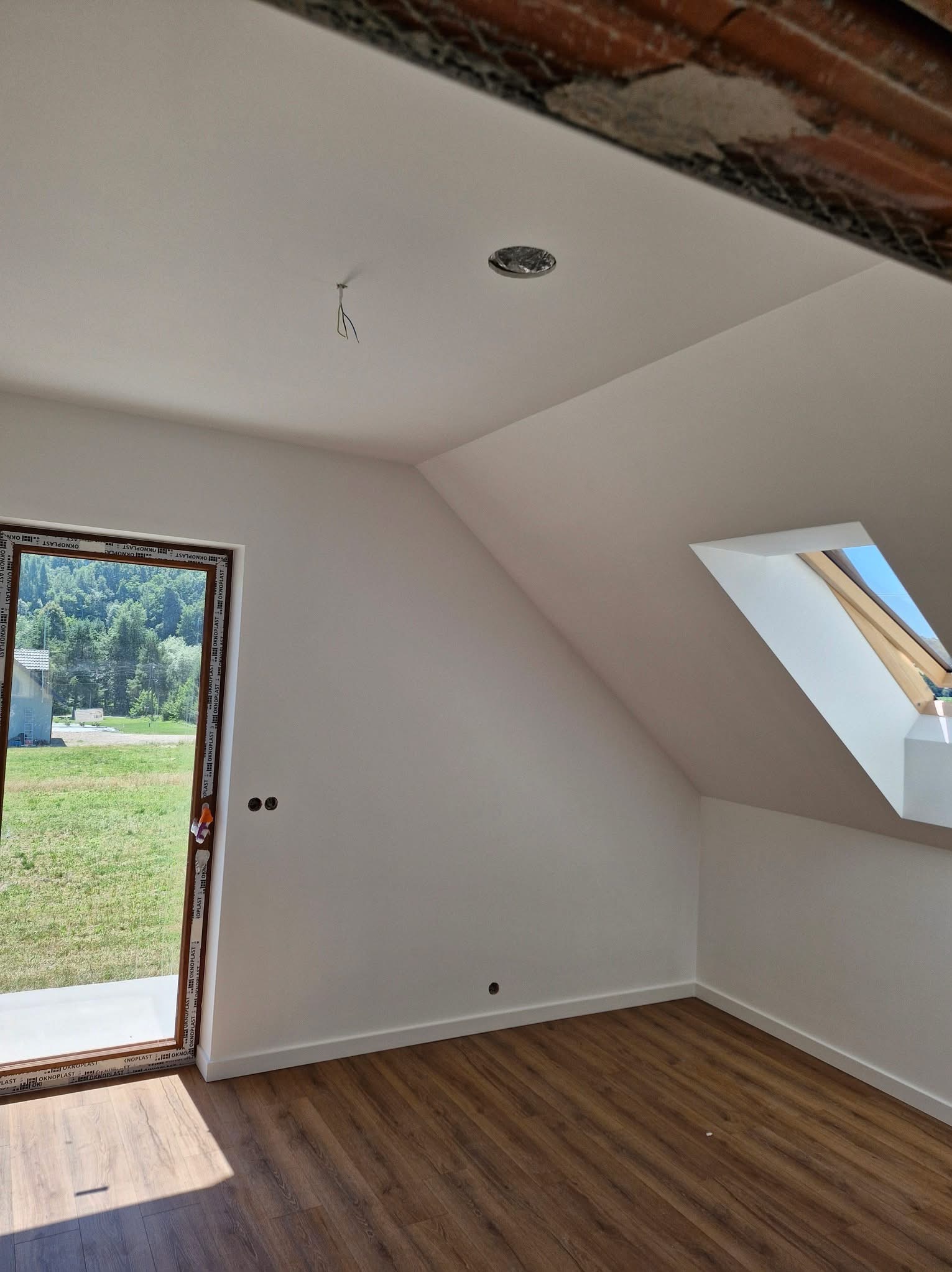 Jasne poddasze po remoncie: białe ściany, drewniana podłoga, okno dachowe i widok na zieleń przez okno balkonowe. Widoczne gniazdka elektryczne i wystające kable.