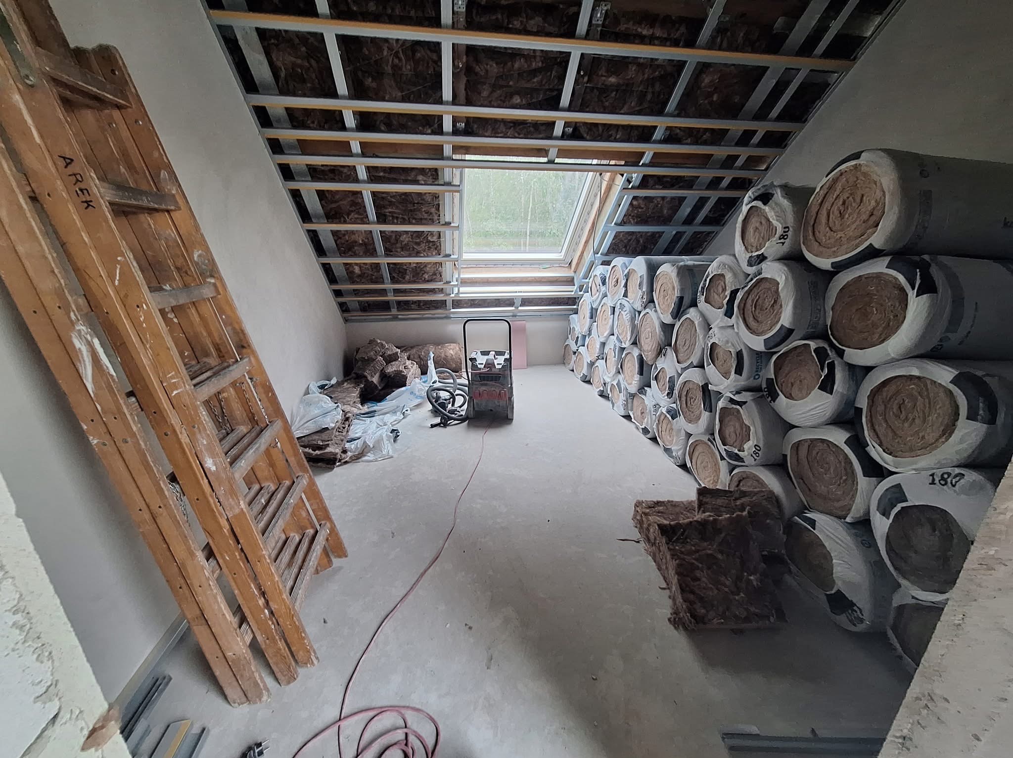 Poddasze w trakcie ocieplania: widoczne rolki wełny mineralnej, metalowy stelaż pod płytę gipsową i okno dachowe. Na podłodze leżą resztki materiału izolacyjnego.