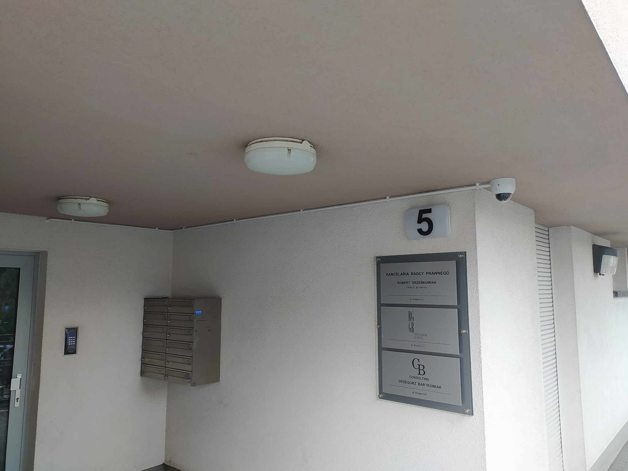 Wejście do budynku z kamerą monitoringu, skrzynkami pocztowymi i tabliczką informacyjną kancelarii radcy prawnego. Widoczne oświetlenie zewnętrzne.