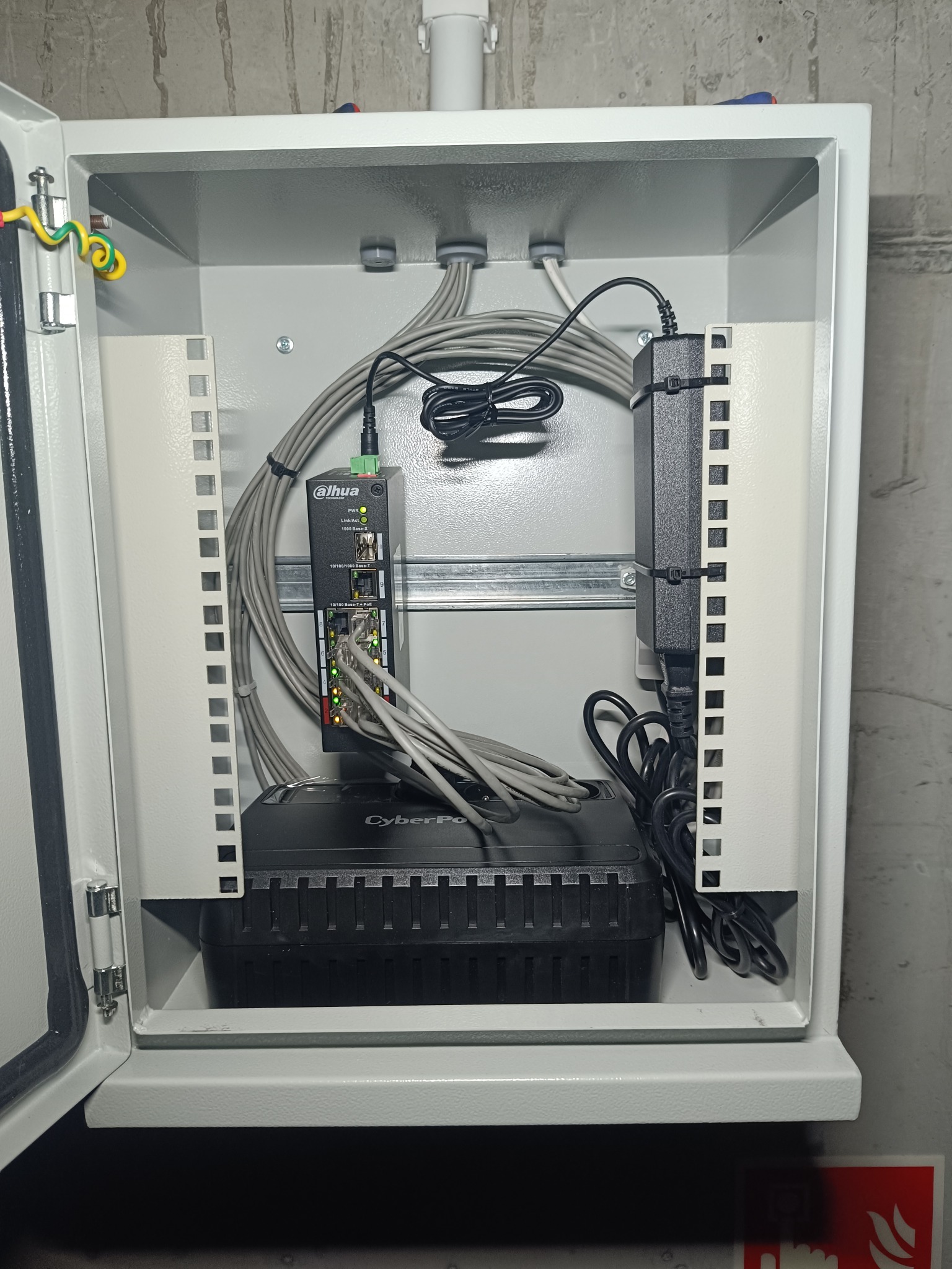 Otwarta metalowa skrzynka z zamontowanym switchem Dahua, zasilaczem i UPS CyberPower. Widoczne kable i przewody. Instalacja sieciowa w szarej obudowie.