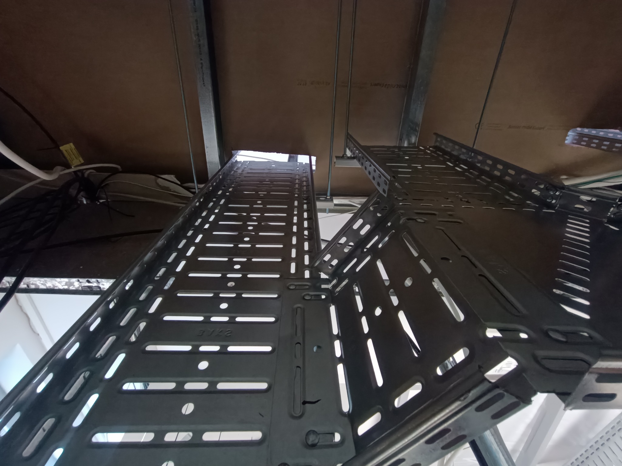 Metalowe korytka kablowe pod sufitem, widok z dołu. Korytka z perforacją, ułożone wzdłużnie, z widocznymi przewodami i elementami konstrukcyjnymi sufitu.