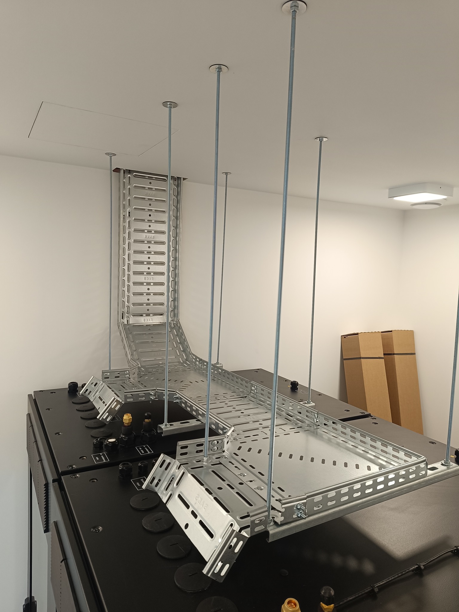 Metalowa trasa kablowa podwieszona do sufitu na prętach, z widocznymi elementami montażowymi i fragmentem szafy rack, w jasnym pomieszczeniu.