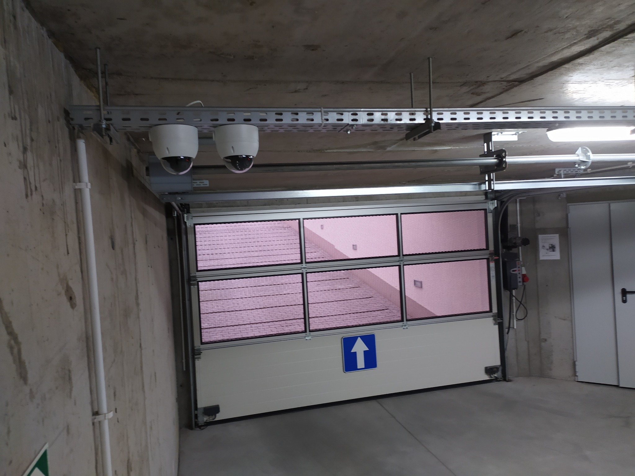 Dwie kamery kopułkowe zamontowane na szynie pod sufitem garażu. Brama segmentowa z oknami i znakiem strzałki w górę. Szare betonowe ściany i podłoga.