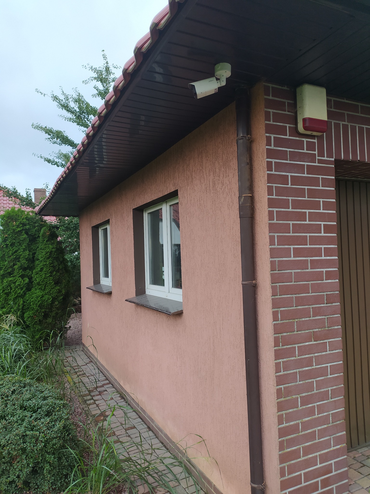Kamera monitoringu i sygnalizator alarmowy zamontowane na elewacji budynku z ceglanym narożnikiem i dwoma oknami. Widok z zewnątrz, obok roślinność.