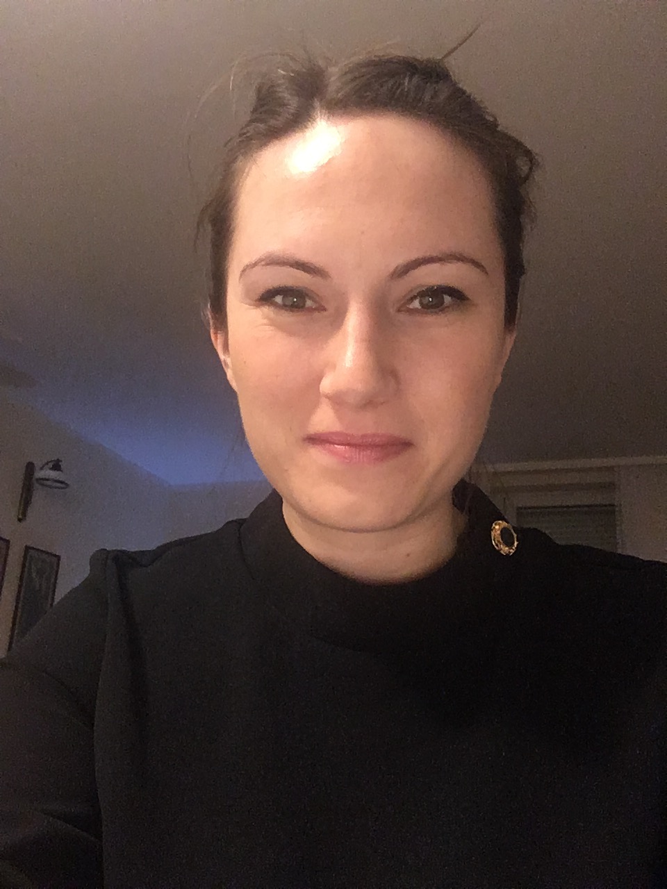 Portret kobiety w czarnej bluzce z ozdobnym złotym guzikiem, uśmiechająca się, z upiętymi włosami. Zdjęcie wykonane w pomieszczeniu.