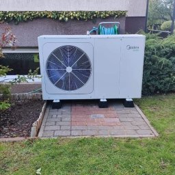 Murawski & Ekorozwiązania-Pro Sp.zo.o - Biała pompa ciepła Midea na kostce brukowej w ogrodzie, z zielonym wężem na górze. Widoczne rośliny i fragment elewacji budynku.