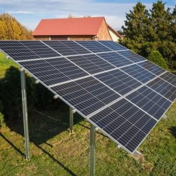 Murawski & Ekorozwiązania-Pro Sp.zo.o - Instalacja paneli fotowoltaicznych na zielonej posesji. Widoczny montaż na metalowych wspornikach, w tle budynek z czerwonym dachem i drzewa.