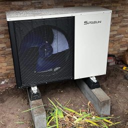 Murawski & Ekorozwiązania-Pro Sp.zo.o - Biała pompa ciepła SPRSUN z czarnym wentylatorem, zamontowana na betonowych blokach przy ścianie z kamienia dekoracyjnego. Widoczne narzędzia i ścięte rośliny.