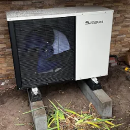 Biała pompa ciepła SPRSUN z czarnym wentylatorem, zamontowana na betonowych blokach przy ścianie z kamienia dekoracyjnego. Widoczne narzędzia i ścięte rośliny.