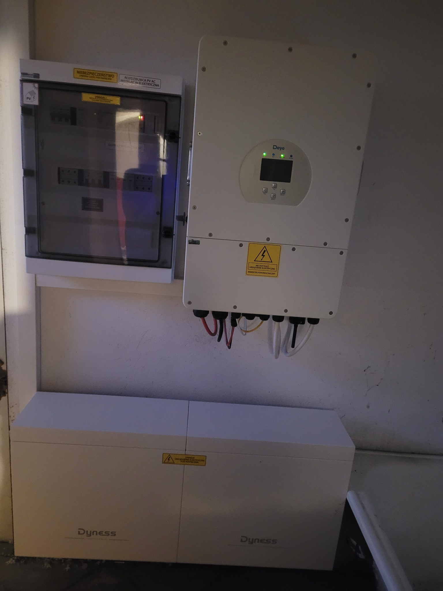 Kompletna instalacja fotowoltaiczna: rozdzielnica, falownik Deye z diodami i akumulator Dyness. Elementy połączone przewodami, widoczne naklejki ostrzegawcze.