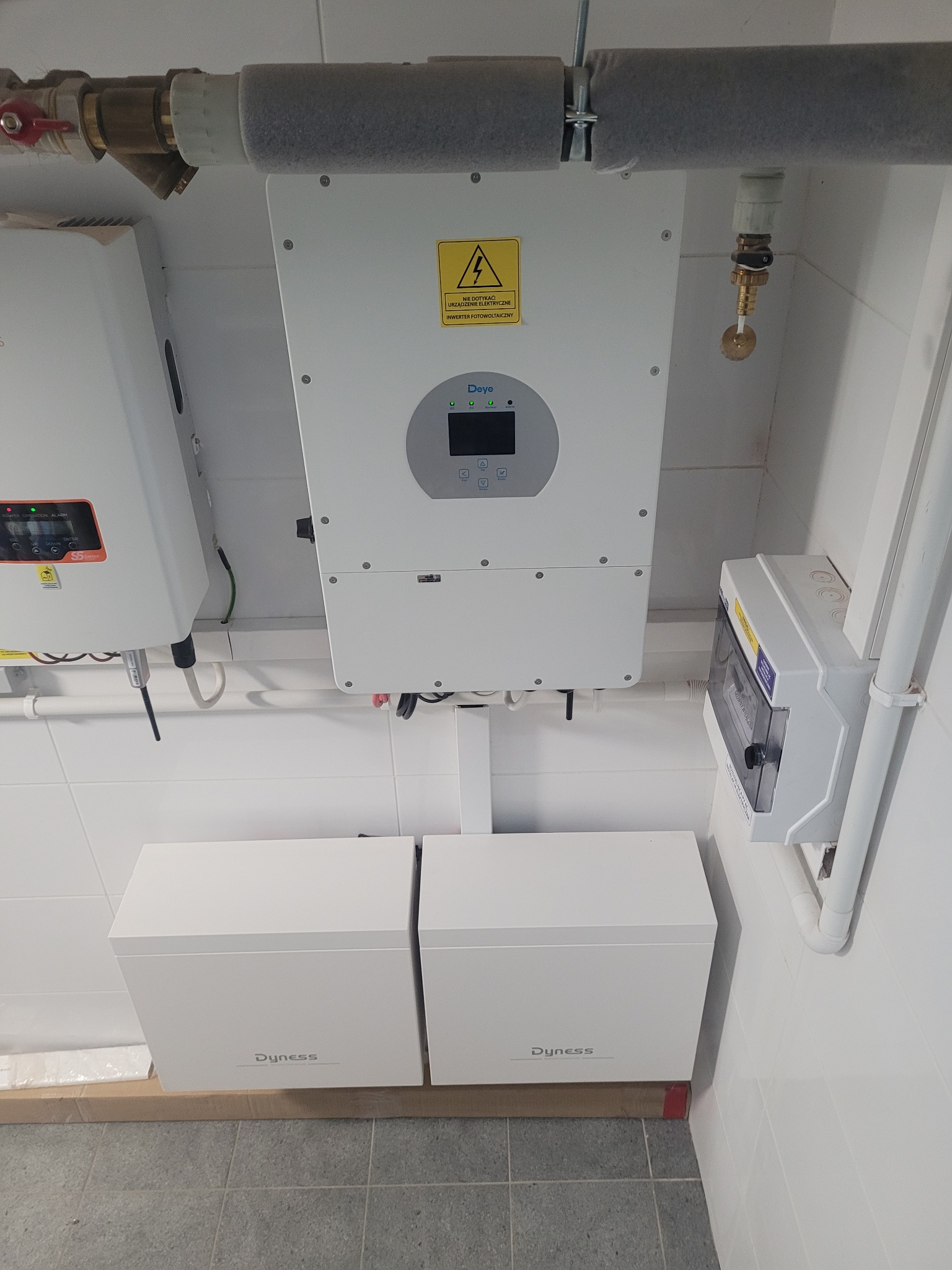 Białe magazyny energii Dyness pod inwerterem Deye, zamontowane na kafelkowanej ścianie. Widoczne rury instalacyjne i skrzynka elektryczna. Szare płytki podłogowe.