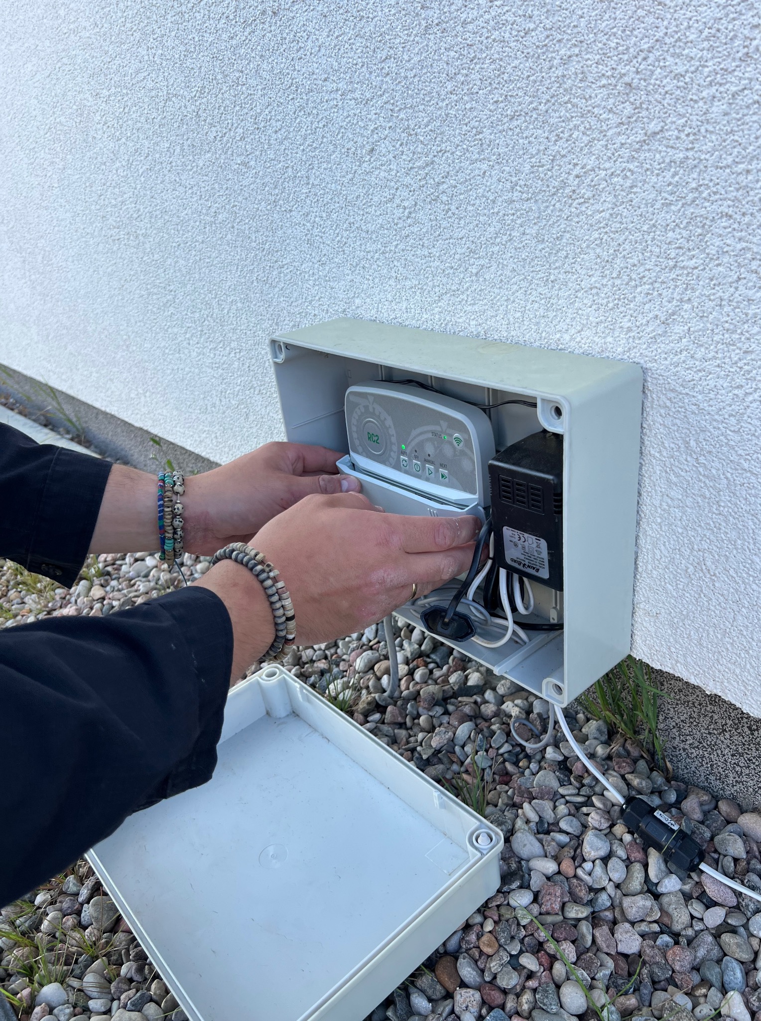Instalacja sterownika nawadniania Rain Bird w białej obudowie na elewacji budynku. Widoczne dłonie montera i elementy instalacji elektrycznej.