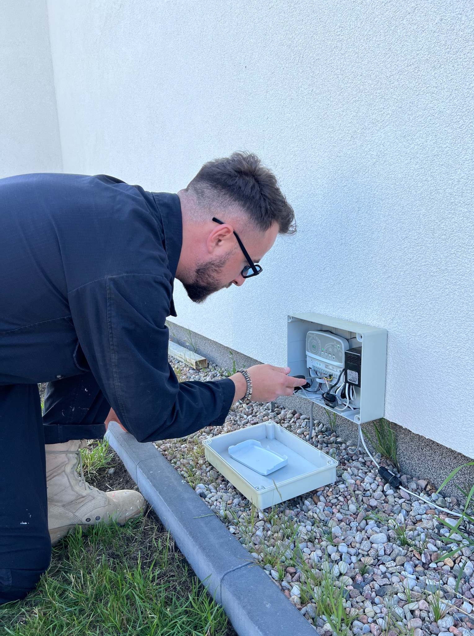 Instalator w kombinezonie podłącza sterownik systemu nawadniania w szarej obudowie na elewacji budynku. Obok leży biała kuweta na narzędzia.