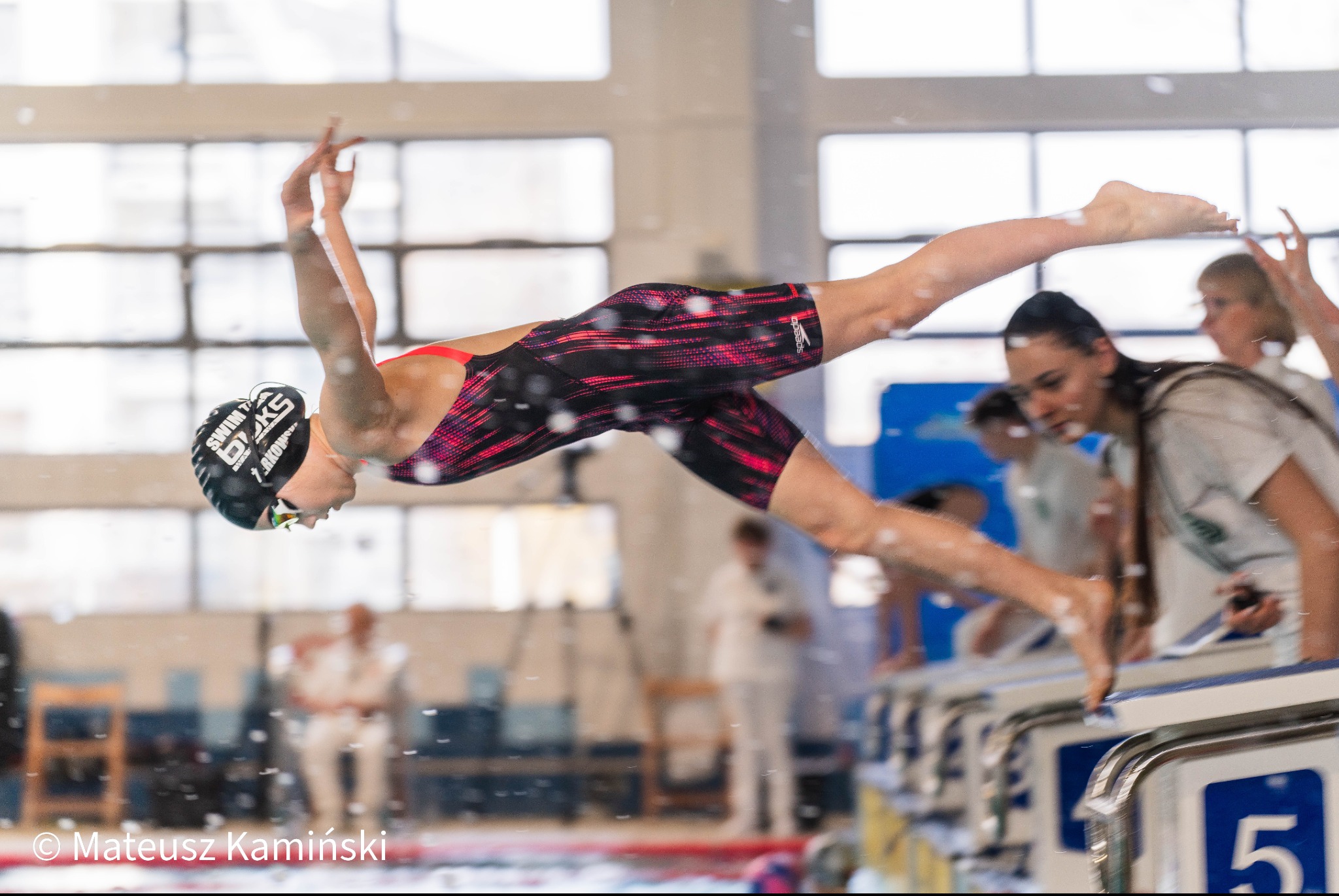 Zawodniczka w locie nurkuje do basenu ze słupka startowego, z czepkiem i okularami, w tle inni zawodnicy i okna. Dynamiczne ujęcie skoku do wody.