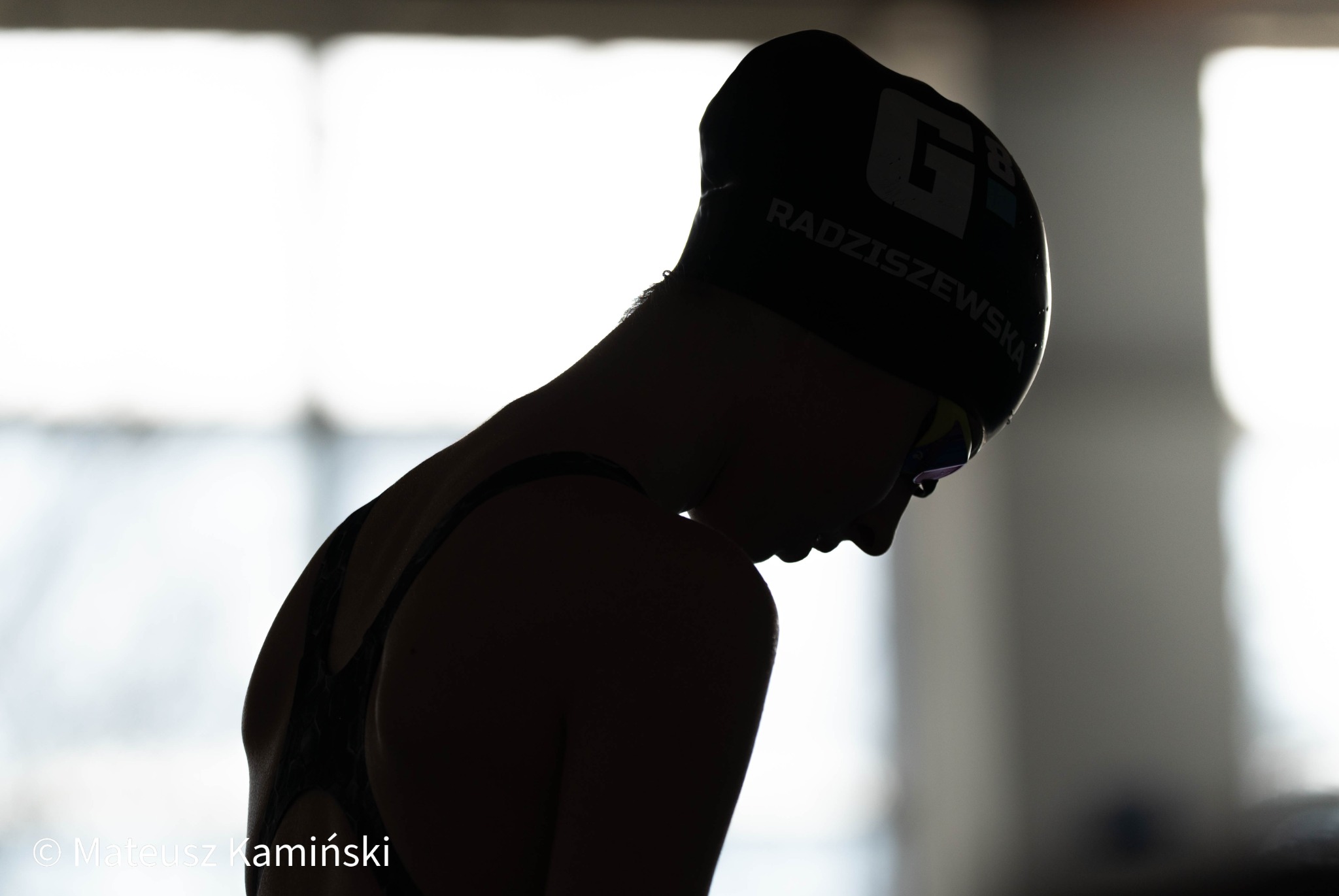 Sylwetka młodej pływaczki w czepku z napisem, pochylona głowa, widoczne okulary pływackie na tle jasnego okna. Przygotowanie do treningu pływackiego.