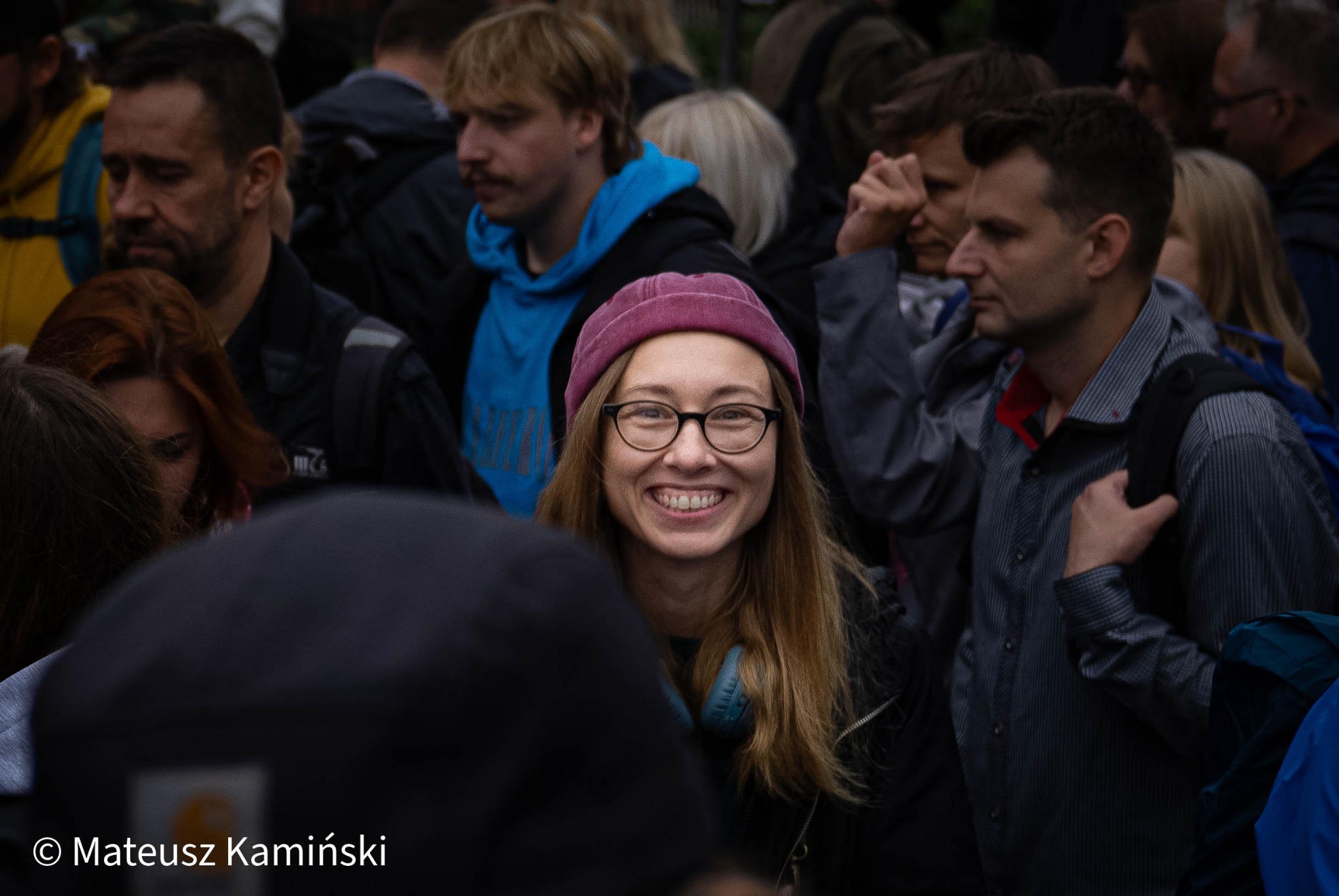 Uśmiechnięta kobieta w okularach i bordowej czapce, otoczona przez tłum ludzi. Widoczne słuchawki na szyi. Zdjęcie z wydarzenia plenerowego z naturalnym oświetleniem.