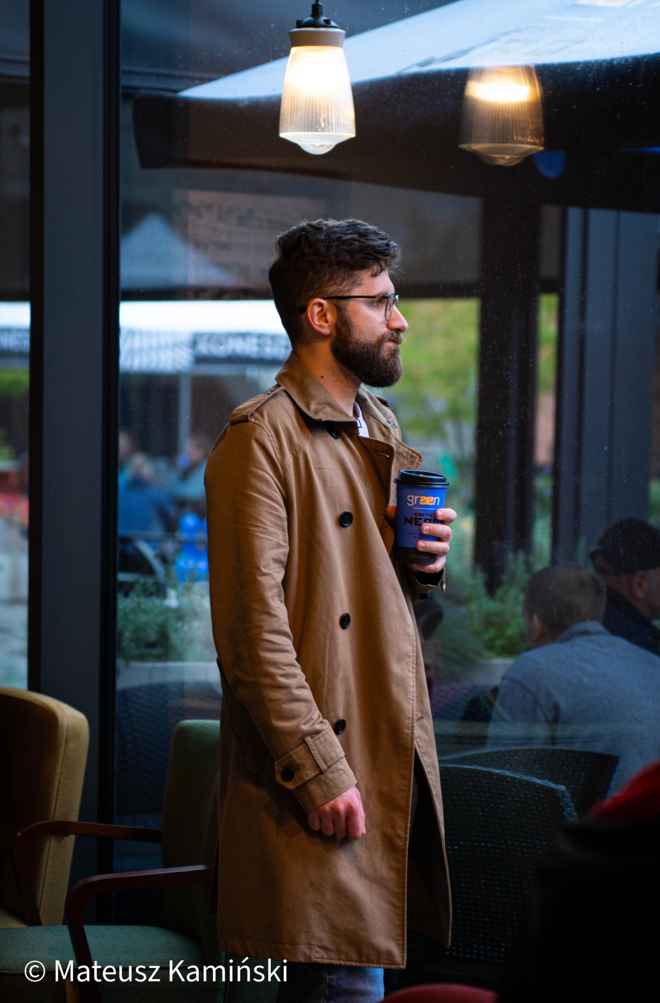 Mężczyzna w płaszczu trzyma kubek kawy w kawiarni. Profil, widoczne odbicia w szybie, lampa sufitowa. Neutralne kolory, naturalne światło.