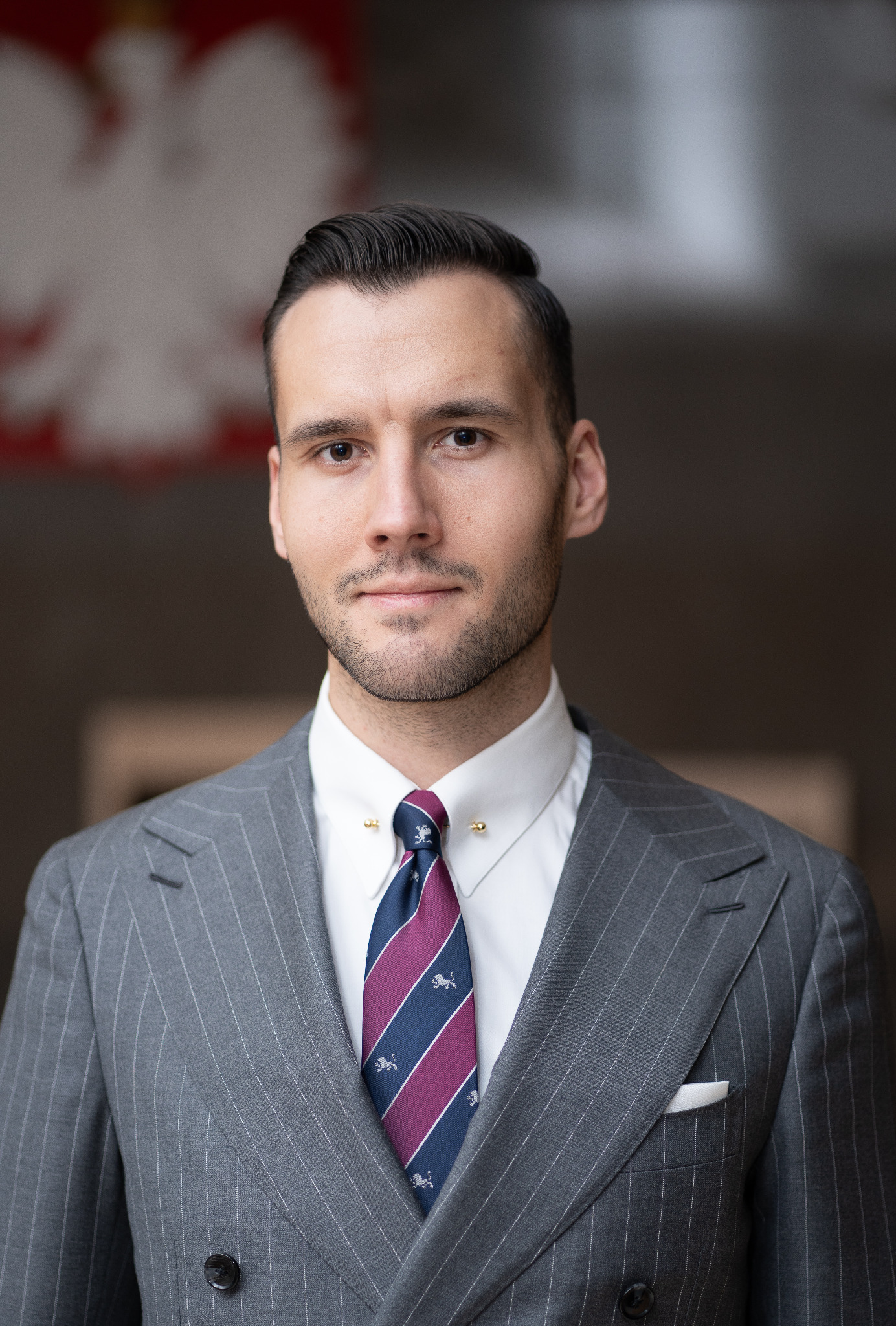 Portret mężczyzny w garniturze w prążki, białej koszuli i krawacie z motywem lwa na tle rozmytego godła Polski.