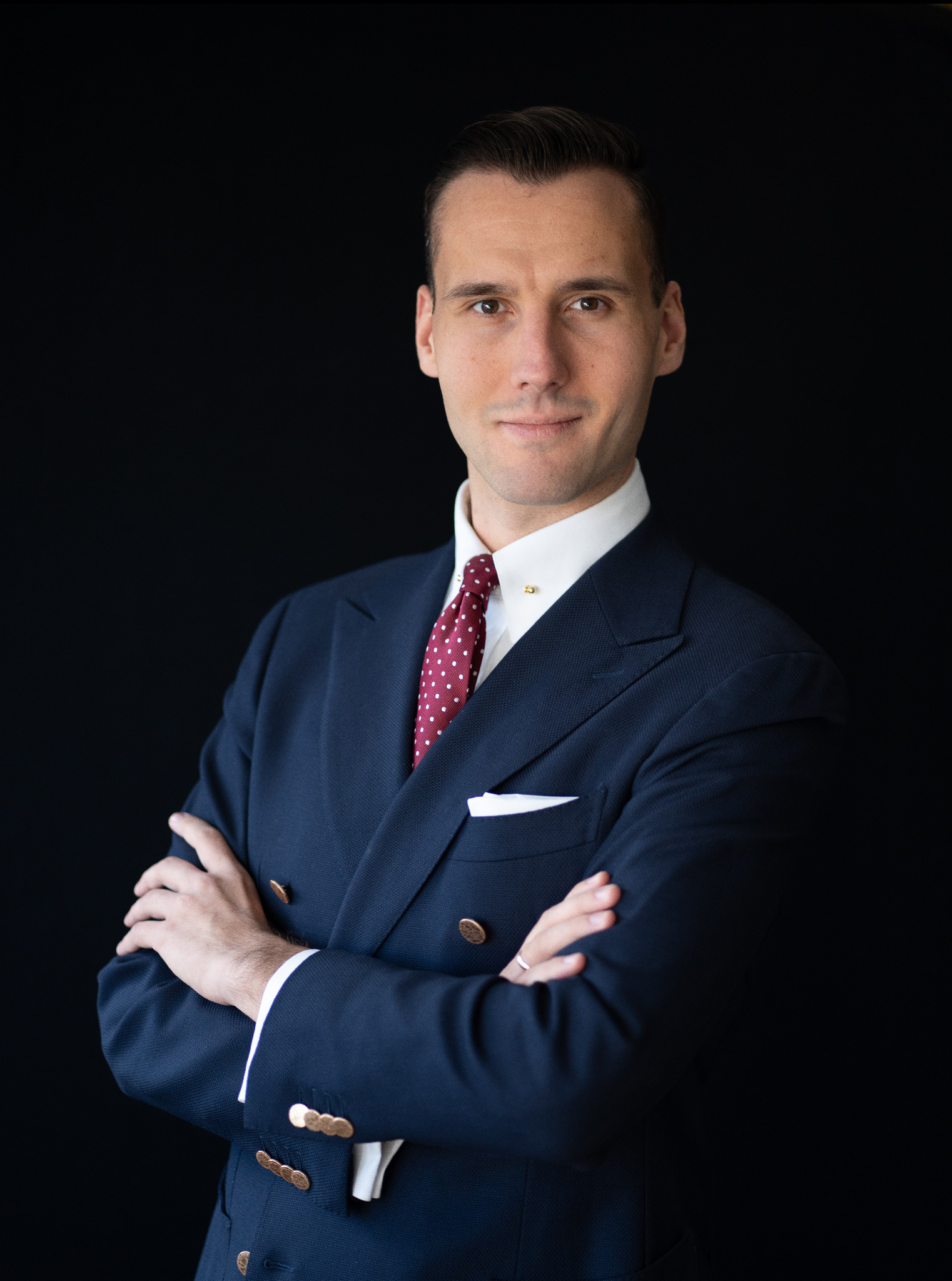 Portret pewnego mężczyzny w granatowym garniturze i bordowym krawacie w kropki, z rękami skrzyżowanymi na tle czerni, sugerujący profesjonalizm i zaufanie.