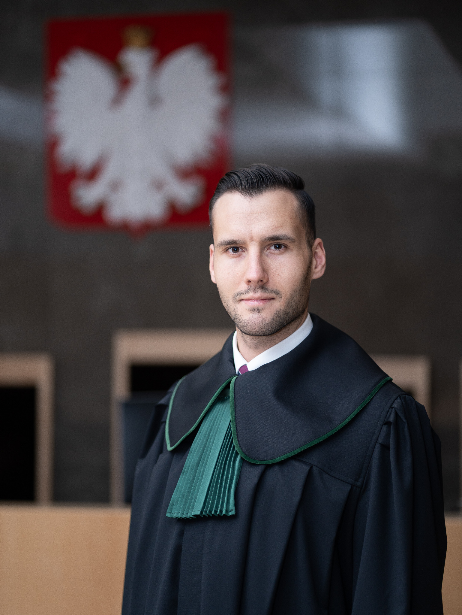 Młody adwokat w todze na tle godła Polski w sądzie. Profesjonalny portret prawnika, skupiony wzrok, ciemne włosy. Ujęcie od pasa w górę, neutralne tło.