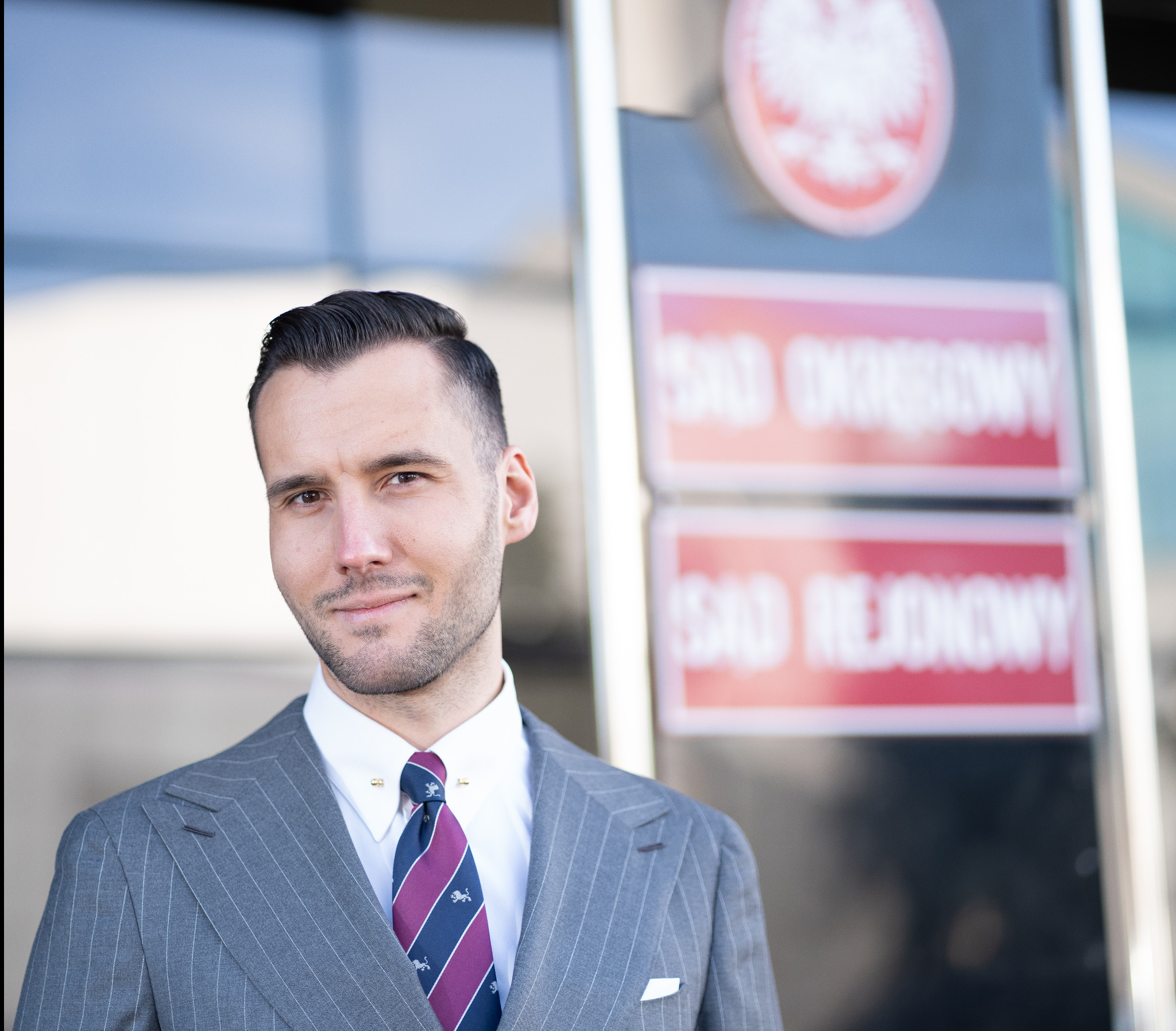 Portret adwokata w garniturze na tle Sądu Okręgowego i Rejonowego. Elegancki strój, pewny wyraz twarzy. Krawat w paski z motywem lwa.