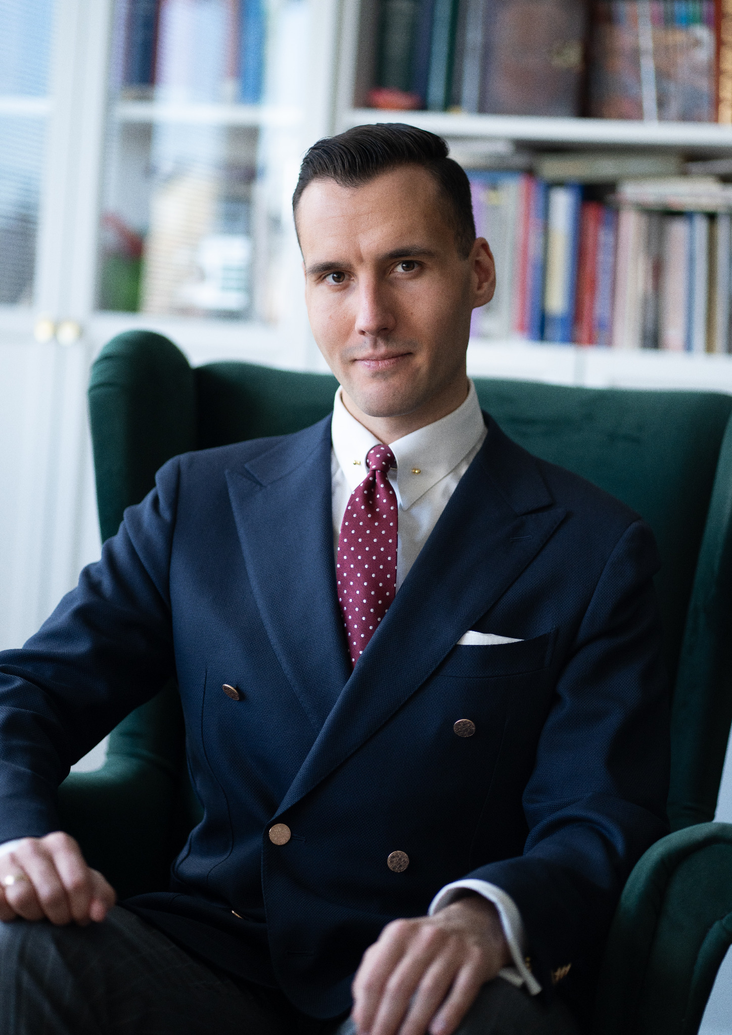 Portret mężczyzny w eleganckim, dwurzędowym garniturze, siedzącego w zielonym fotelu na tle regału z książkami. Wyraźny krawat w kropki i spinka.